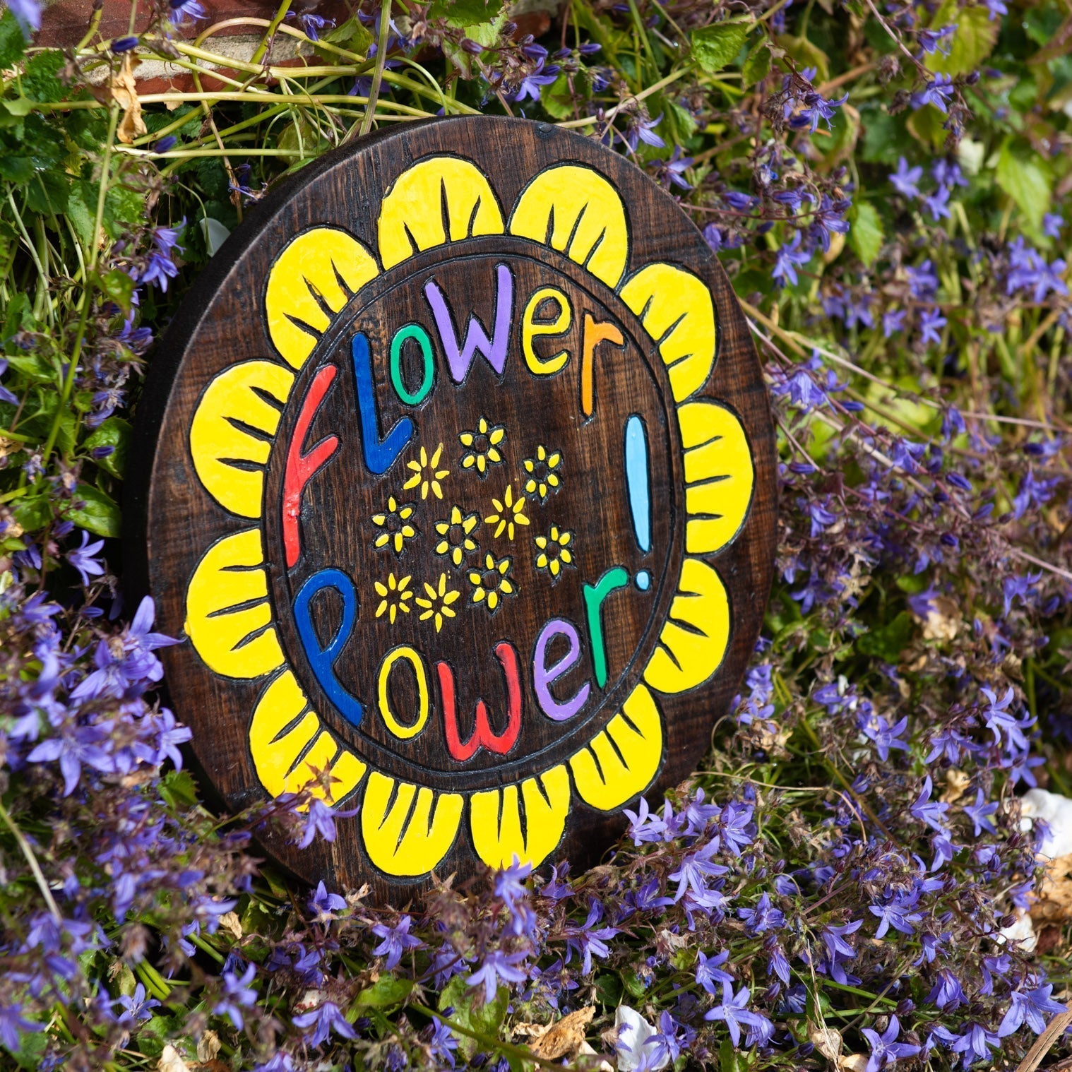 Flower Power wood plaque propped in a bed of blue flowers, yellow petals and bold painted text on dark stained disc