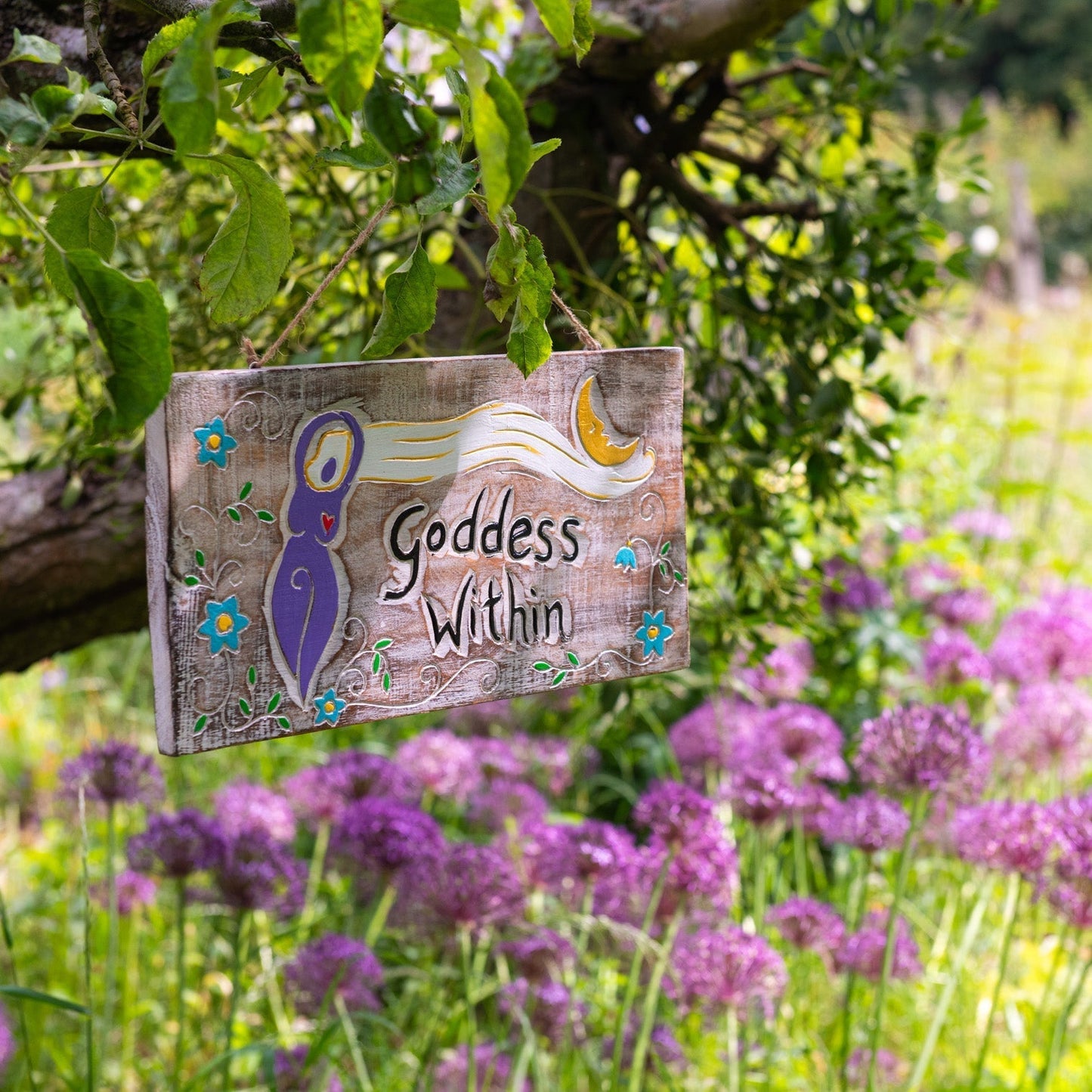 Carved wood Goddess Within plaque hanging from a tree branch, showing purple goddess figure, crescent moon and blue flowers