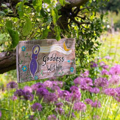 Carved wood Goddess Within plaque hanging from a tree branch, showing purple goddess figure, crescent moon and blue flowers