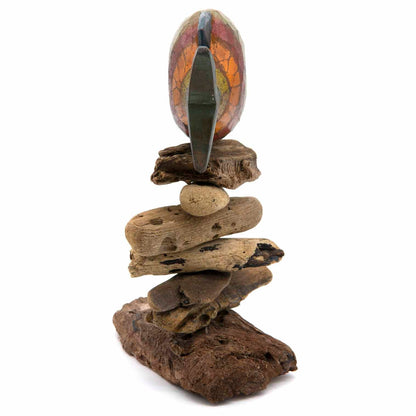 Painted oval wood carving in orange and red on driftwood and pebble stack, side view