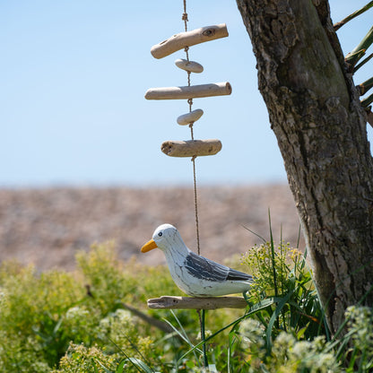Hanging wooden seagull with driftwood sticks and pebbles on jute cord, outdoors against blue sky