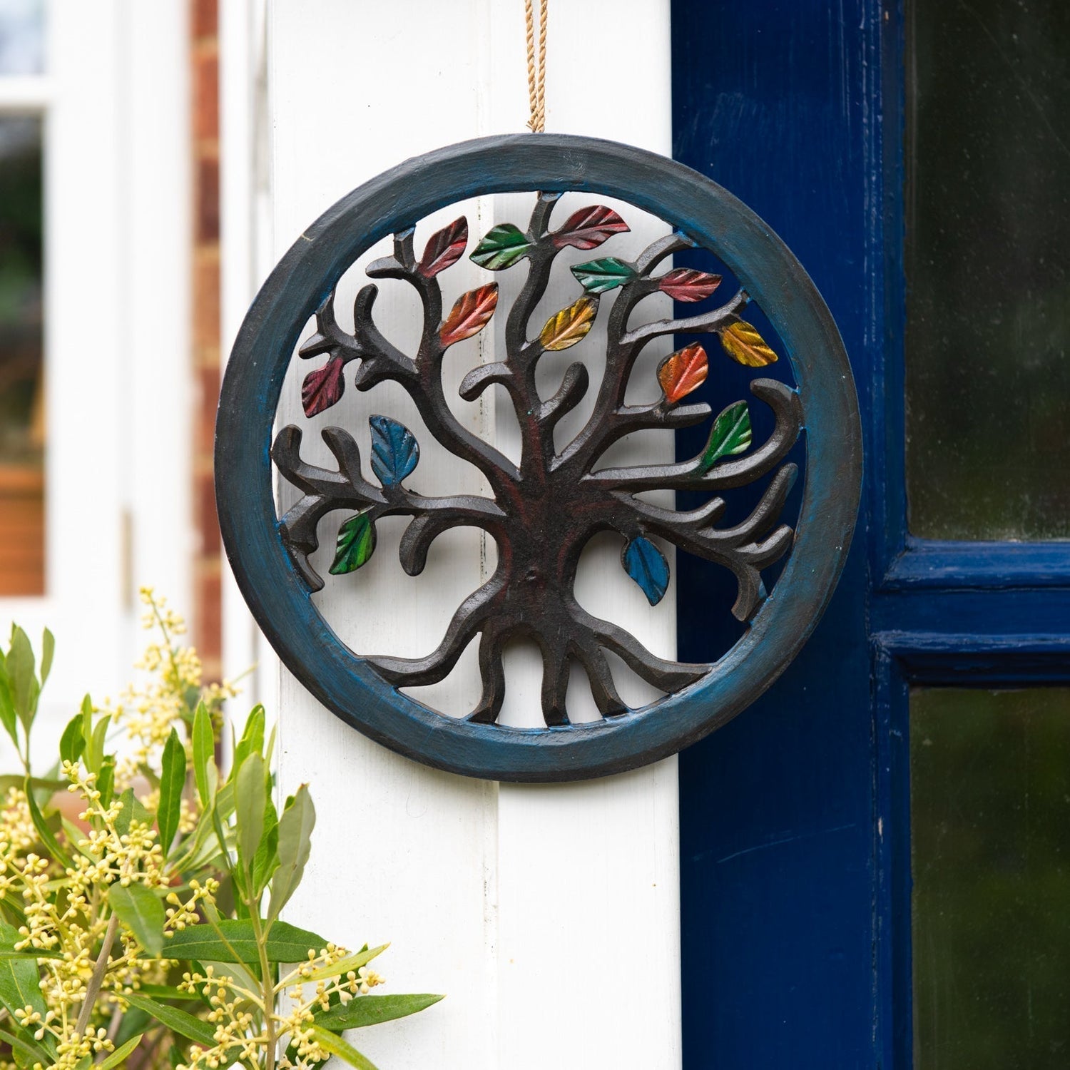 Circular Tree of Life wood plaque with autumn-coloured leaves hanging on a white-painted exterior wall beside a blue door