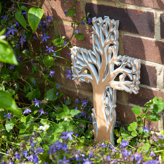Whitewash Tree of Life cross plaque leaning against a brick wall among bluebells, showing pale finish against warm wood