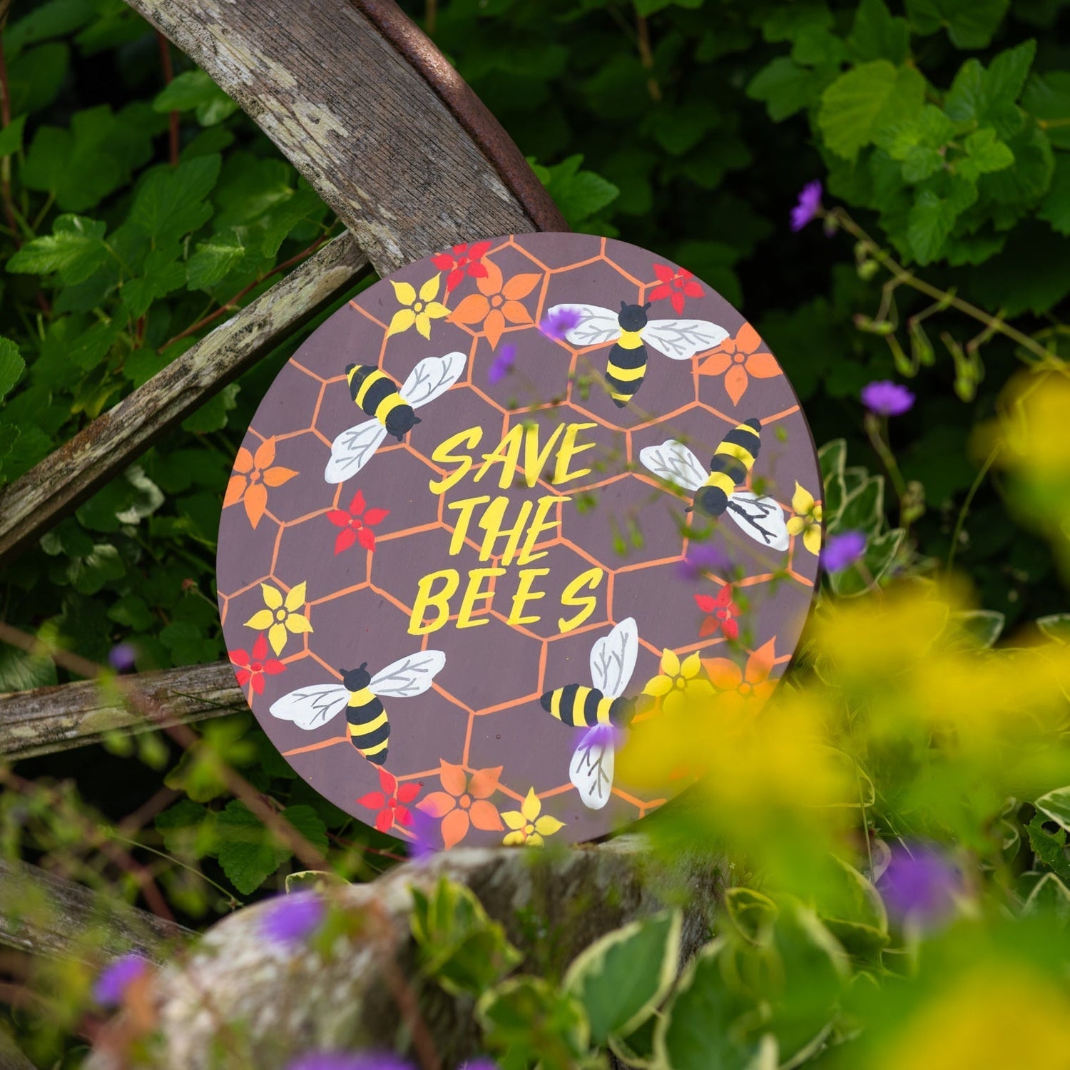 Round bee plaque with honeycomb pattern and yellow lettering hanging on a rustic wooden fence among wildflowers
