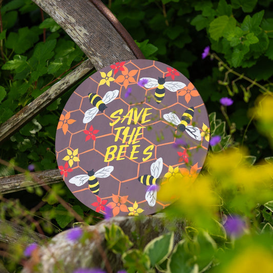 Round bee plaque with honeycomb pattern and yellow lettering hanging on a rustic wooden fence among wildflowers