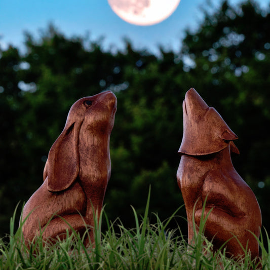 Large amber wood howling wolf and moon gazing hare carvings outdoors under a full moon