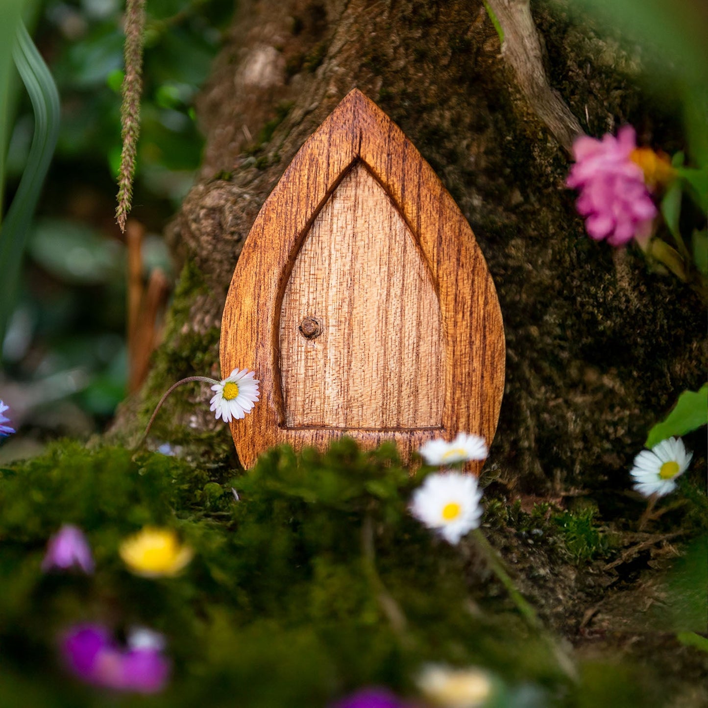 Carved wooden fairy door with pointed arch set against a mossy tree trunk among flowers