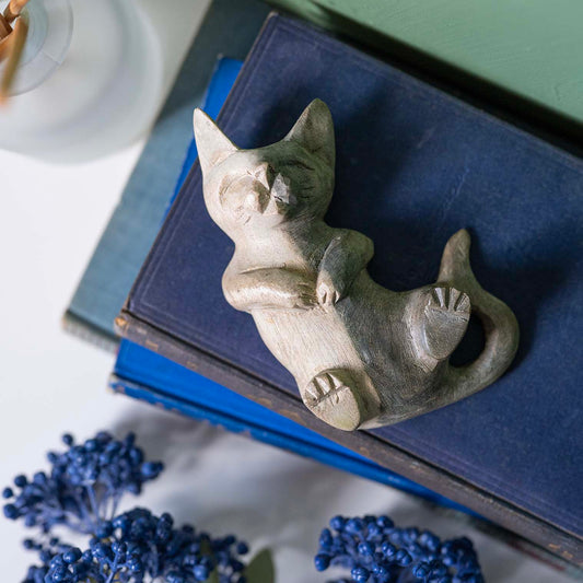 Hand-carved wooden cat on its back with paws raised, pale Indonesian wood with bleached finish, displayed on navy book