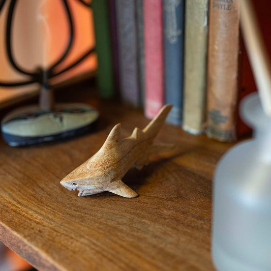 Hand-carved wooden shark with open jaw and spread fins, resting on a wooden desk beside magnifying glass and craft books
