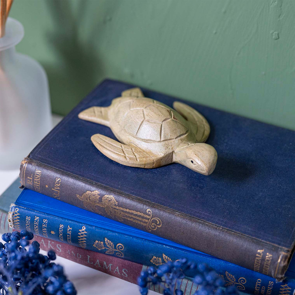 Hand-carved pale wood sea turtle with faceted hexagonal shell and crosshatched flippers, resting on stacked vintage books
