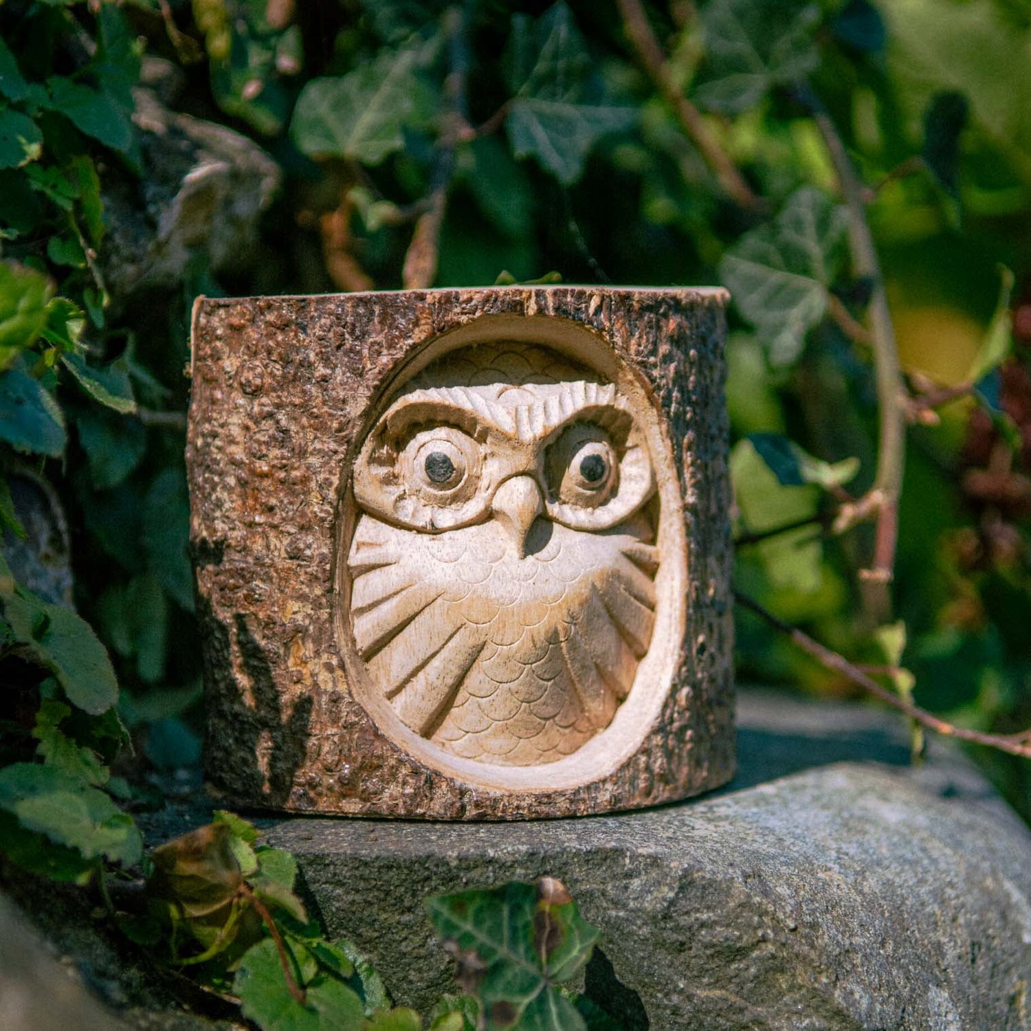 Owl face carved in relief into a natural log section with bark surround, front three-quarter view outdoors among ivy, Indones