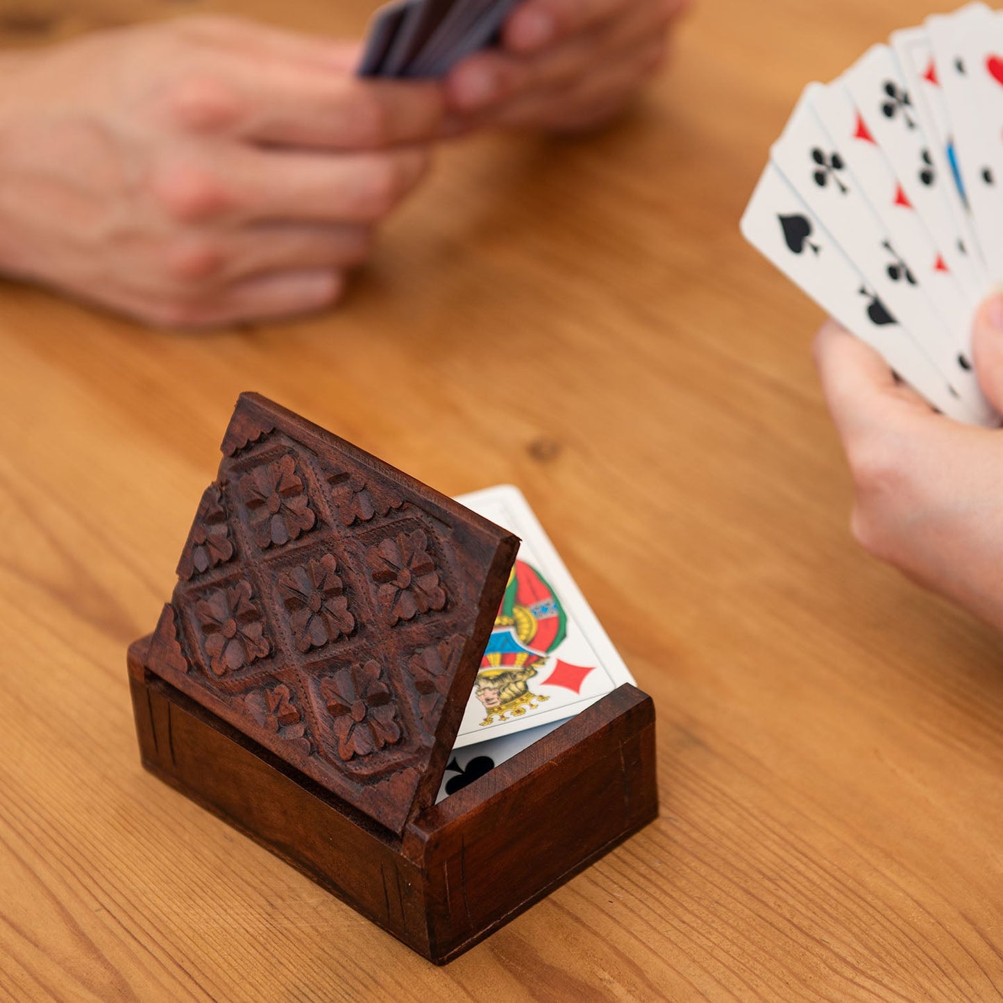 Carved wood playing card box propped open with a deck of cards inside, on a wooden table during a card game
