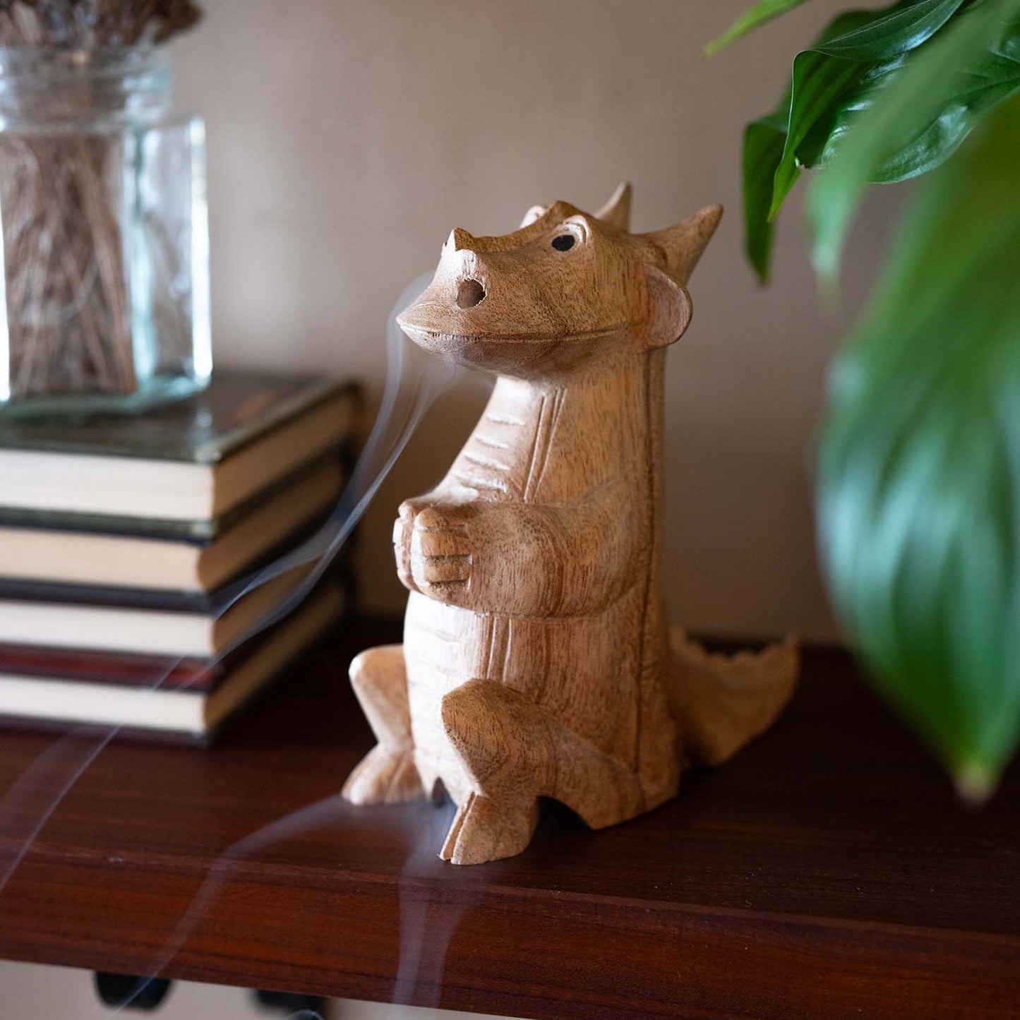 Hand-carved wooden smoking dragon with smoke curling from nostrils, seated upright on wooden shelf surrounded by books and greenery