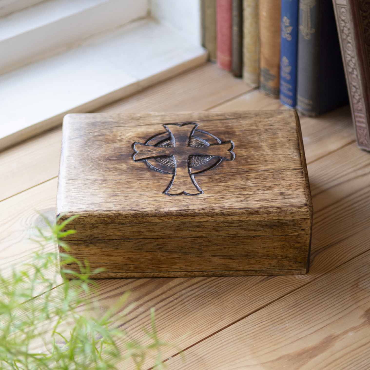 Mango wood box with engraved Celtic cross lid, resting on wooden floor beside books and plant
