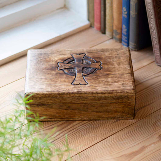 Mango wood box with engraved Celtic cross lid, resting on wooden floor beside books and plant