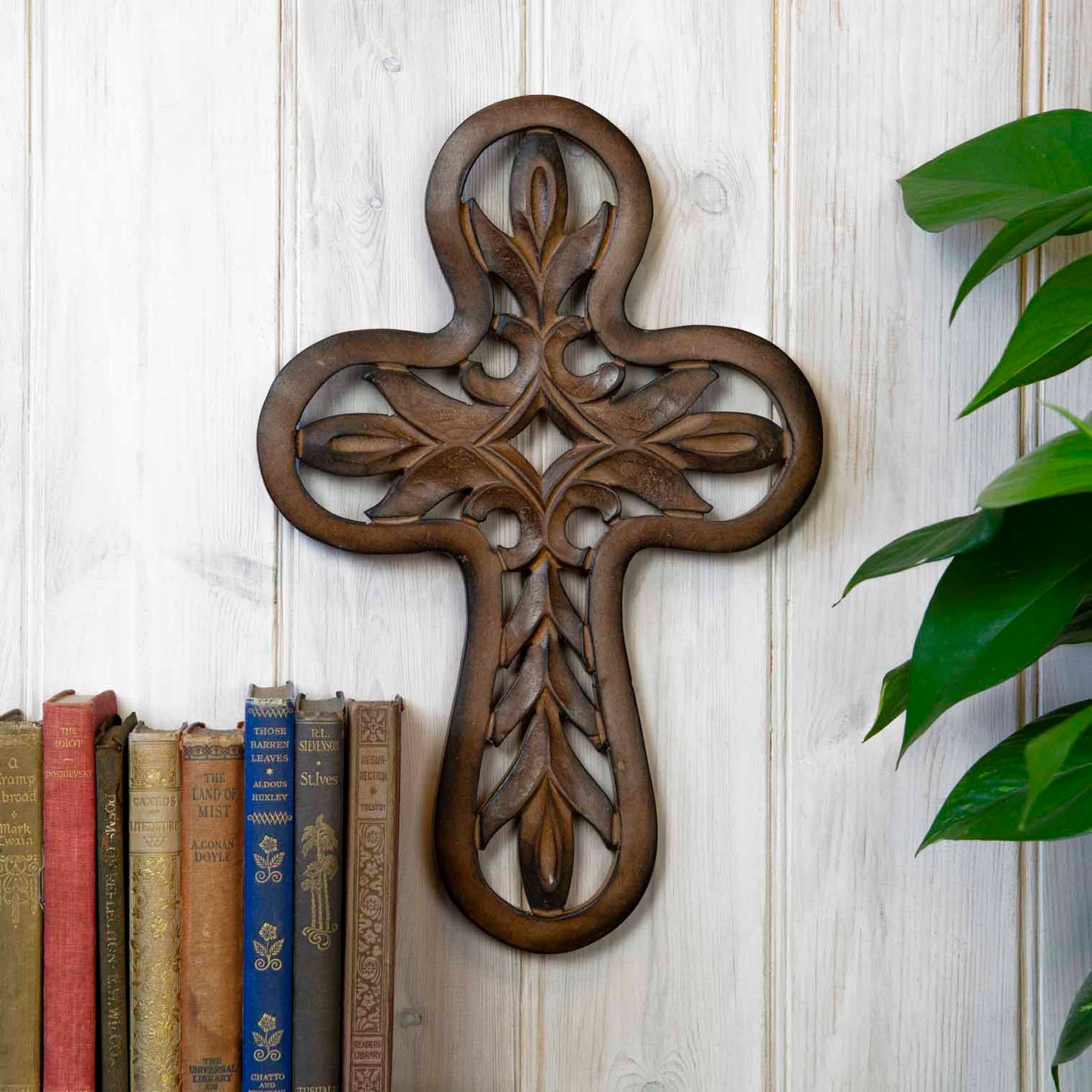 Carved mango wood cross plaque with open leaf lattice, hung on a white-painted wall beside vintage books, India