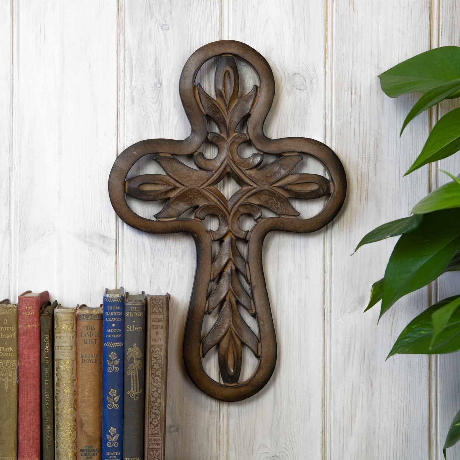 Carved mango wood cross plaque with open leaf lattice, hung on a white-painted wall beside vintage books, India