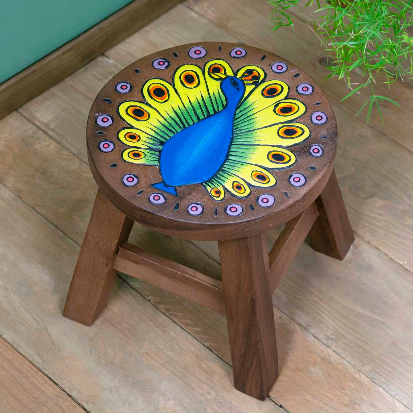 Raintree wood peacock stool with carved blue peacock and fanned green and yellow tail, angled view on wooden floor, Thailand
