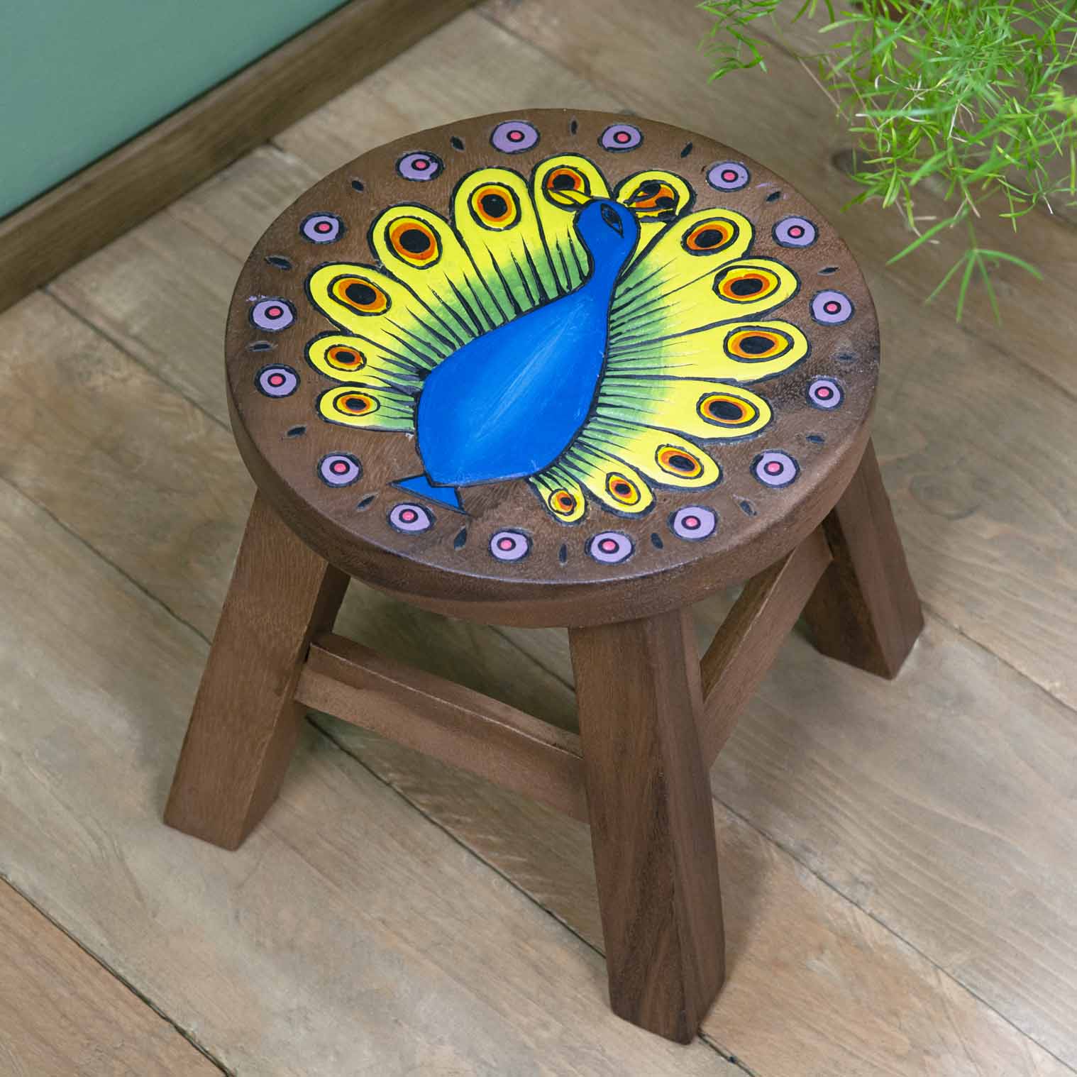 Raintree wood peacock stool with carved blue peacock and fanned green and yellow tail, angled view on wooden floor, Thailand
