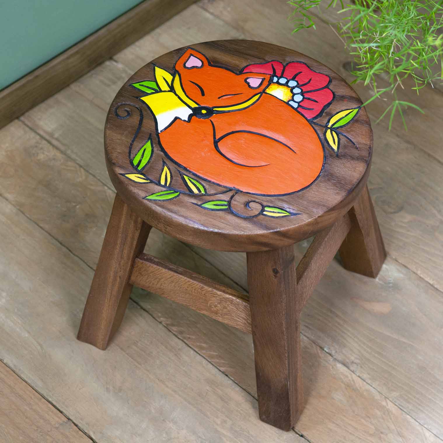 Raintree wood sleeping fox stool with carved orange fox ringed by green leaves and red poppy, angled view on wooden floor
