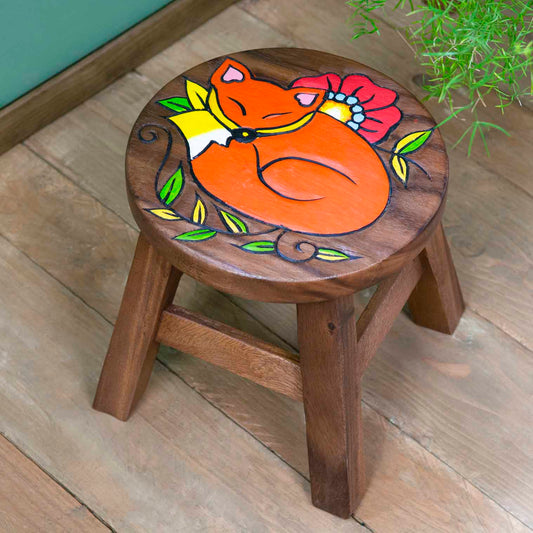 Raintree wood sleeping fox stool with carved orange fox ringed by green leaves and red poppy, angled view on wooden floor